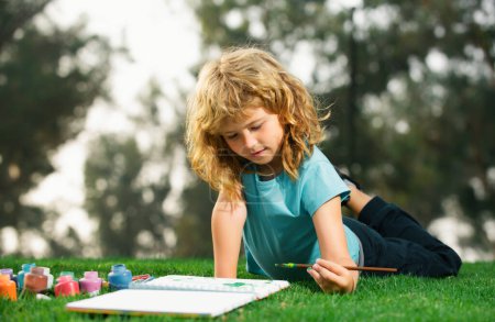 Photo for Kid draws in park laying in grass having fun on nature background. Children artist paints creativity vacation, kids crafts - Royalty Free Image