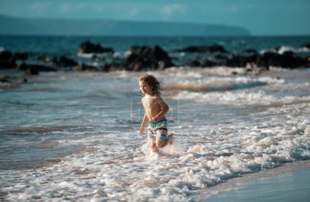 Foto de Un niño feliz corriendo en la playa tropical. Niño jugando olas de mar en el mar de vacaciones en las vacaciones de verano. Niño en la naturaleza en la orilla del mar. Niños caminando en la playa - Imagen libre de derechos