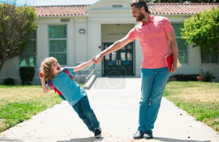 Parent and funny pupil of primary school go hand in hand. Teacher in t-shirt and cute schoolboy with backpack near school park