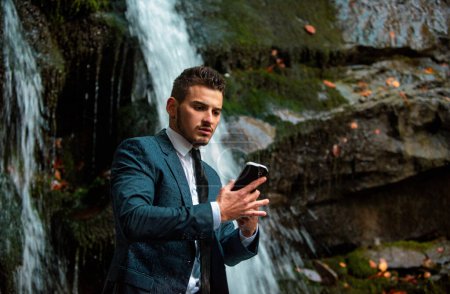 Téléchargez les photos : Homme d'affaires en costume avec smartphone à fond de cascade. Concept d'appareil étanche. Télétravail homme d'affaires avec téléphone ip68 - en image libre de droit