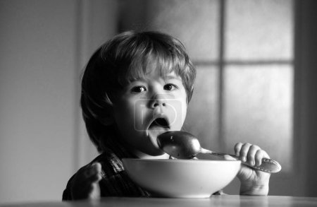 Foto de Niño sentado en la mesa comiendo con expresión divertida en la cara. Sabroso desayuno para niños. Sabroso desayuno para niños. Alegre bebé niño come comida sí mismo con cuchara - Imagen libre de derechos