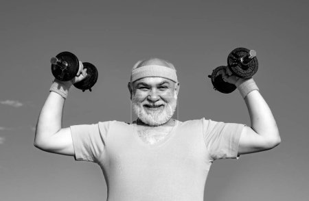 Photo for Happy senior man with dumbbell looking at camera. Body care and healthcare. Grandfather sportsman portrait on blue sky backgrounds - Royalty Free Image