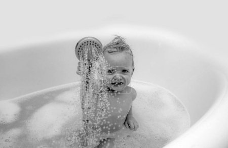 Foto de Un niño sonriente tomando un baño con jabón. Lindo niño tomando baño jugando con espuma y juguetes de pato en un baño blanco y soleado - Imagen libre de derechos
