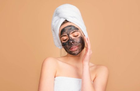 Woman with with charcoal mask touching her nose. Mud facial mask, face clay mask spa. Beautiful woman with cosmetic mud facial procedure, spa health concept. Skin care beauty treatment. Towel on head