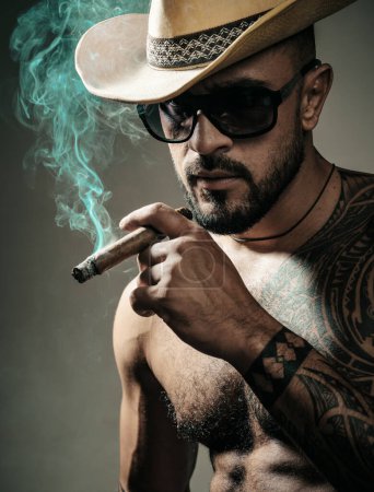 Photo for Seductive face of a sexy man smoking cigar. Macho looking confident. Tattooed and elegant hipster - Royalty Free Image