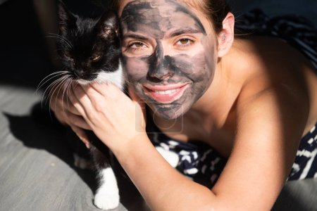Téléchargez les photos : Bonjour avec le chat noir. Masque visage au charbon. Cosmétiques, cosmétologie, dermatologie. Visage de femme avec masque en argile noire. Procédure cosmétique femmes visage dans le masque sur le visage - en image libre de droit