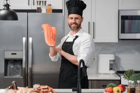 Photo for Chef in cook apron and chef hat at modern kitchen preparing fish salmon. Middle aged man is cooking fresh fish salmon seafood crab and shrimp and lobster in the kitchen at home - Royalty Free Image