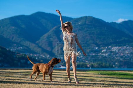Foto de Despreocupado Chica jugar con el perro Boxer cerca de la naturaleza con fondo de montaña. Chica lanzando pelota al perro boxeador. El perro intenta atrapar la pelota. Sensual mujer jugando con el perro en el parque en el día de verano. Hermosa mascota - Imagen libre de derechos