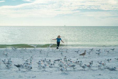 Photo for Carefree kid running, chasing birds. Kid boy chasing birds near summer sea beach. Happy child playing with seagull birds outside on summer day - Royalty Free Image