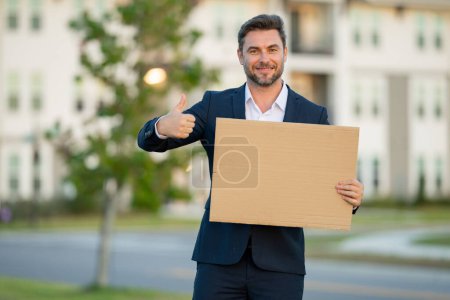 Photo for Businessman presenting signboard, billboard or banner. Business man with blank banner ad, posing outdoor. Portrait of attractive business man in business suit holding empty with thumb up - Royalty Free Image