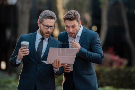 Two surprised businessman using laptop outdoor. Handsome business man in suits working on laptop and communication with business projects in city. Partners thinking and plan for business
