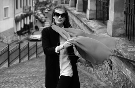 Téléchargez les photos : Photo de style de rue d'une élégante femme à la mode portant des vêtements à la mode avec une écharpe soufflant dans le vent. Modèle marchant dans la rue de la ville européenne. Foulard venteux - en image libre de droit