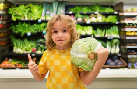 Téléchargez les photos : Enfant au chou. Enfant au supermarché aux légumes. Petit enfant qui choisit de la nourriture en magasin ou à l'épicerie - en image libre de droit