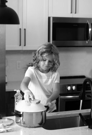 Téléchargez les photos : Enfant garçon laver la vaisselle dans l'évier de la cuisine. Nettoyage vaisselle pendant les travaux ménagers - en image libre de droit