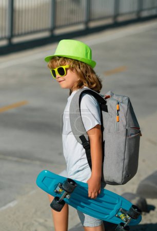 Téléchargez les photos : Joyeux enfant élégant dans les lunettes de soleil et chapeau d'été de mode posant avec planche à roulettes en plein air. Sports d'été en plein air et mode de vie - en image libre de droit
