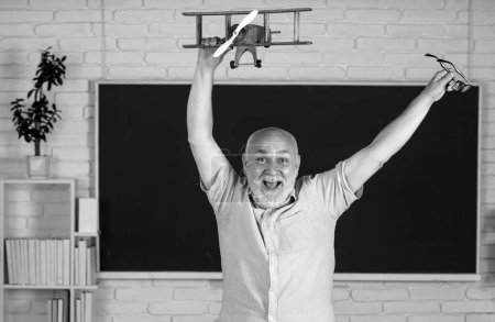 Photo for Portrait of excited senior teacher teaching line of high school students with computer laptop in classroom on blackboard. Amazed aged male retired teacher with toy plane - Royalty Free Image