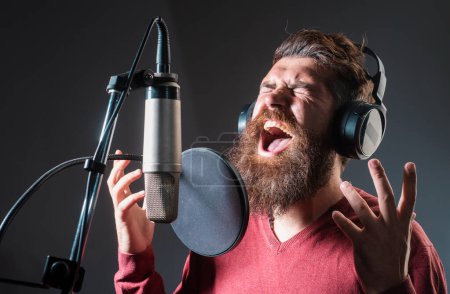 Photo for Singer man wearing headphones is performing a song with a microphone while recording in a music studio - Royalty Free Image