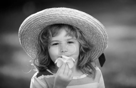 Téléchargez les photos : Portrait d'un mignon enfant garçon en chapeau de paille sentant la fleur de plumeria. Gros plan sur les enfants caucasiens. Gros plan tête de drôle de gosse - en image libre de droit