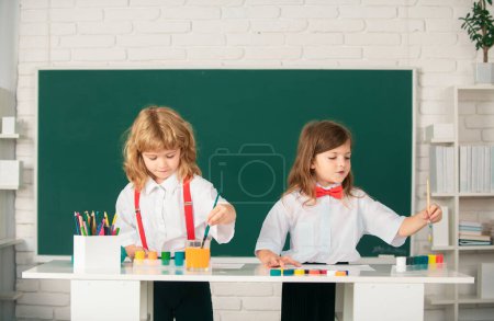 Téléchargez les photos : École enfants fille et garçon peinture avec des peintures couleur et pinceau dans la salle de classe. Enfants mignons dessinent avec des crayons colorés - en image libre de droit