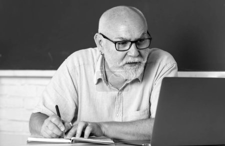 Photo for Elderly education and retired knowledge concept. Senior teacher teaching in the classroom on chalkboard. Teacher at university. Man senior teacher using laptop computer sitting at desk in classroom - Royalty Free Image