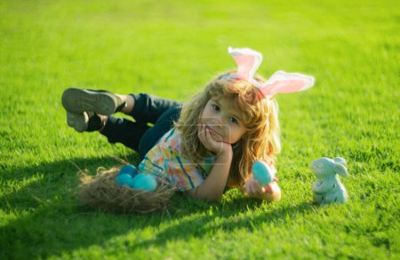 Bunny boy hunting eggs in park, Funny little Easter bunny child hunt easter eggs laying on grass in backyard. Easter kids portrait outdoor