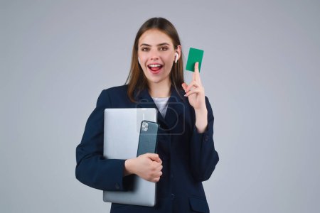 Foto de Mujer joven en camiseta blanca con tarjeta de crédito cerca de la cara y el ordenador portátil, aislado sobre fondo gris. Paga con tarjeta de crédito o débito. Tarjeta bancaria. Tarjeta de crédito de plástico - Imagen libre de derechos