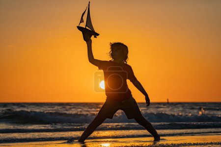 Téléchargez les photos : Silhouette d'enfant jouant avec jouet bateau à voile sur la mer coucher du soleil. Enfant avec bateau jouet dans l'eau de mer en vacances d'été. Petit enfant jouant avec jouet bateau à voile sur la mer. Vacances d'été. Journée des enfants - en image libre de droit