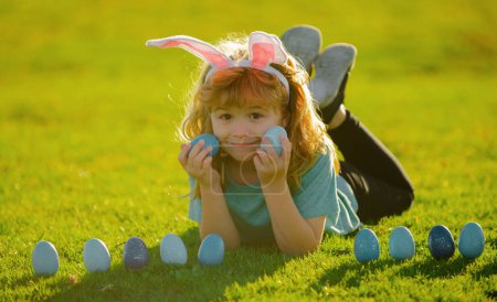 Téléchargez les photos : Les enfants chassent les œufs de Pâques. Concept vacances printemps enfants. Enfant garçon avec des œufs de Pâques et des oreilles de lapin sur l'herbe. Lapin de Pâques enfants - en image libre de droit