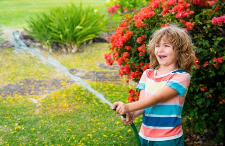 Foto de Lindo niño regando flores en el jardín en el día de verano. Niño usando manguera de jardín. Divertido niño regando plantas en el jardín del patio - Imagen libre de derechos