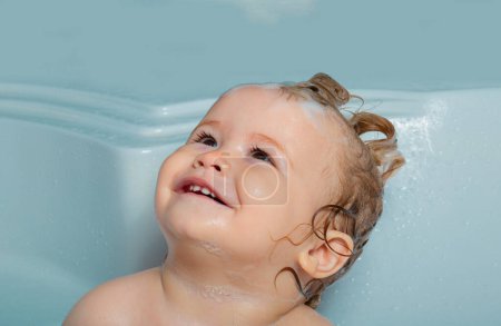 Téléchargez les photos : Joyeux visage de bébé dans le bain moussant. Bains de bébé heureux drôles dans la baignoire avec de l'eau et de la mousse. Hygiène des enfants - en image libre de droit