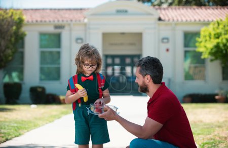 Téléchargez les photos : Un écolier qui va à l'école avec son père. Un petit écolier qui mange à l'extérieur. Déjeuner scolaire pour enfant - en image libre de droit