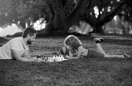 Téléchargez les photos : Fils étendu sur l'herbe et jouant aux échecs avec son père. Enfant apprenant à jouer aux échecs. Petit garçon pense ou planifie le jeu d'échecs. Échec mat - en image libre de droit