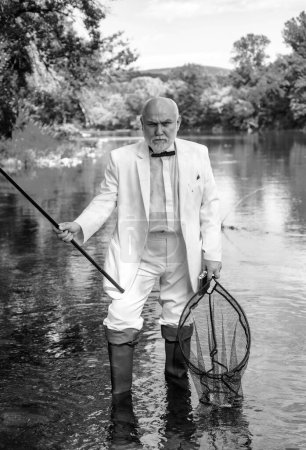 Photo for Fishing hobby and summer weekend. Portrait of senior businessman fishing. Mature man fisherman in white suit and bowtie with fishing rod, spinning reel on river - Royalty Free Image