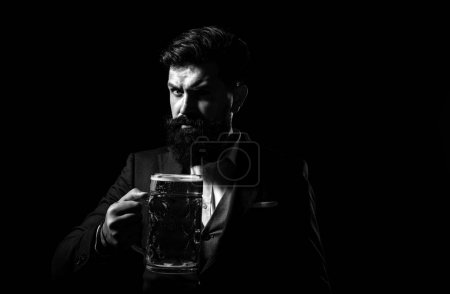 Serious man in classic suit drinking beer. Bearded guy in business outfit looks happy and satisfied. Portrait of man with lifted high glass of beer on black background