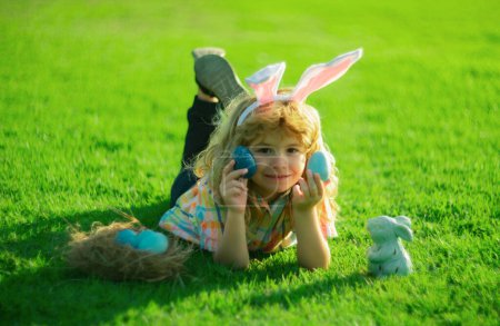 Téléchargez les photos : Lapin de Pâques enfants avec des oreilles de lapin dans le jardin chasse oeuf. Les œufs de chasse d'enfant portent des oreilles de lapin tiennent les œufs de Pâques pondus sur l'herbe dans le parc - en image libre de droit