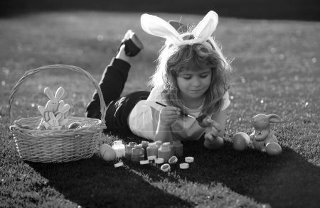 Foto de Niño con huevos de Pascua y orejas de conejo que ponen en huevo de pintura de hierba. Feliz cara de niños de Pascua - Imagen libre de derechos