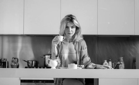 Foto de Mujer sexy beber café cerca de la mesa en la cocina en casa. Sexy modelo que cubre gran reposabrazos con pijama - Imagen libre de derechos