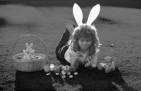 Téléchargez les photos : Enfant garçon en costume de lapin avec des oreilles de lapin peignant des œufs de Pâques sur l'herbe dans le parc de printemps - en image libre de droit
