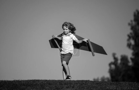Téléchargez les photos : Un gamin jouant avec des ailes en carton. Enfant dans un champ d'été. Les enfants voyagent et vacances concept. Imagination et concept de liberté. Garçon avec des ailes sur le terrain imagine pilote et rêves de voler - en image libre de droit