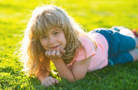Foto de Feliz niñito tirado en la hierba en el día de primavera. Retrato de un niño sonriente acostado sobre hierba verde en el parque. Lindo niño en el prado en el patio trasero - Imagen libre de derechos
