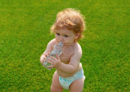 Téléchargez les photos : Le bébé boit l'eau minérale du biberon. Enfant se reposant dans un parc gazonné - en image libre de droit