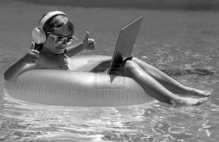 Téléchargez les photos : Les affaires d'été. Enfant travaillant à distance sur ordinateur portable dans la piscine. Petit homme d'affaires travaillant en ligne sur ordinateur portable dans l'eau de la piscine d'été. Bureau d'été et concept freelance - en image libre de droit