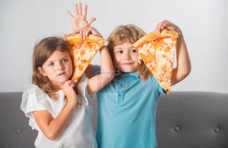 Téléchargez les photos : Les enfants mangent des pizzas. Heureux enfants drôles manger de la pizza et s'amuser ensemble. Enfants drôles tenant une tranche de pizza près du visage - en image libre de droit
