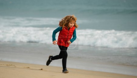 Téléchargez les photos : Joyeux gamin qui court sur la plage. Un drôle de garçon court le long du bord du surf. Mode de vie actif des enfants. Course matinale avec enfants - en image libre de droit