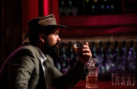 Attractive bearded man in whiskey bar. Hipster with beard and mustache in suit drinks alcohol after working day