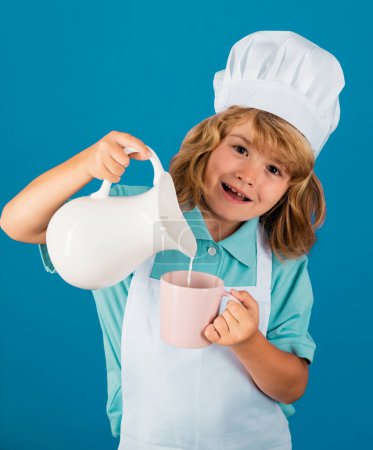 Téléchargez les photos : Portrait d'un enfant de 7, 8 ans en casquette et tablier de cuisine faisant de la salade de fruits et cuisiner des aliments dans la cuisine. Lait pour enfants. - en image libre de droit