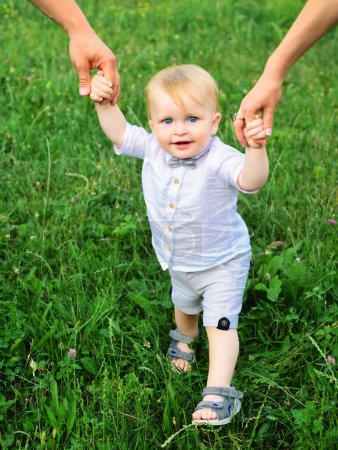 Téléchargez les photos : Les parents main et enfant premier pas. Bébé jouant dans l'herbe verte. Enfant s'amusant sur le pique-nique en famille dans le jardin d'été. Portrait d'une jeune famille heureuse enseignant à bébé à marcher dans le parc - en image libre de droit
