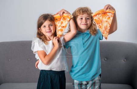 Photo for Happy daughter and son eating pizza. Children kids enjoy and having fun with lunch together at home. Funny children eating pizza and having fun together, holding pizza slice near face - Royalty Free Image