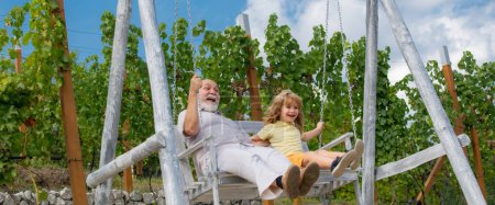 Foto de Nieto hijo y abuelo balanceándose en el jardín de verano. Abuelo y nieto sentados en el columpio en el parque. Abuelo y nieto en el fondo del cielo, bandera de primavera - Imagen libre de derechos