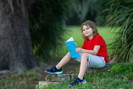 Foto de Lindo niño leyendo un libro en el parque. El chico se sienta en la hierba y lee el libro. Niño leyendo libro de interés en el jardín. Diversión veraniega. Lindo chico leyendo un libro para niños - Imagen libre de derechos
